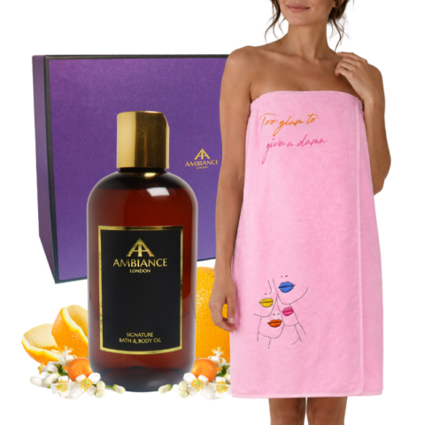 A woman wrapped in a pink towelling wrap dress. A bottle of Ancienne Ambiance London bath oil. The background features a purple gift box and fresh citrus fruits.