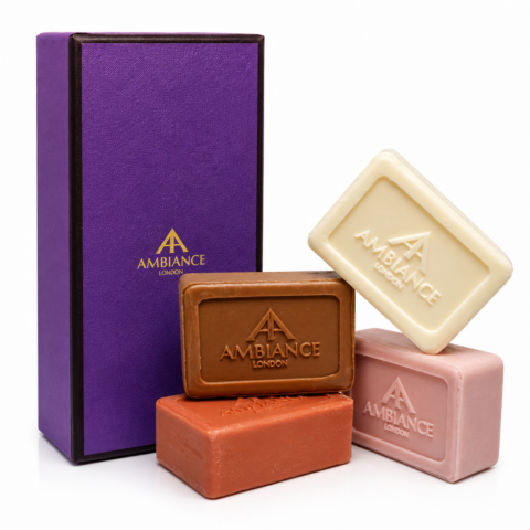 Four bars of soap in ivory, tan, orange, and pink, with "Ancienne Ambiance London" embossed logo. They're arranged beside an elegant purple box.
