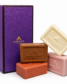 Four bars of soap in ivory, tan, orange, and pink, with "Ancienne Ambiance London" embossed logo. They're arranged beside an elegant purple box.