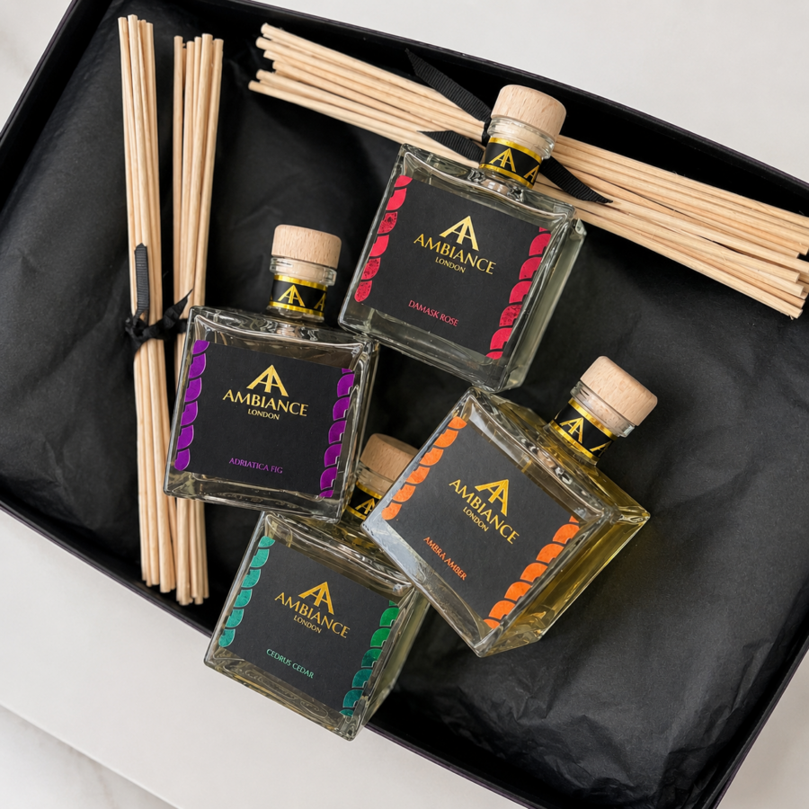 A gift box contains four square reed diffuser bottles labeled "Ancienne Ambiance London" in different scents: Damask Rose, Adriatica Fig, Ambra Amber, and Cedrus Cedar. Natural diffuser sticks are neatly placed beside them on black tissue paper. The arrangement conveys elegance and luxury.