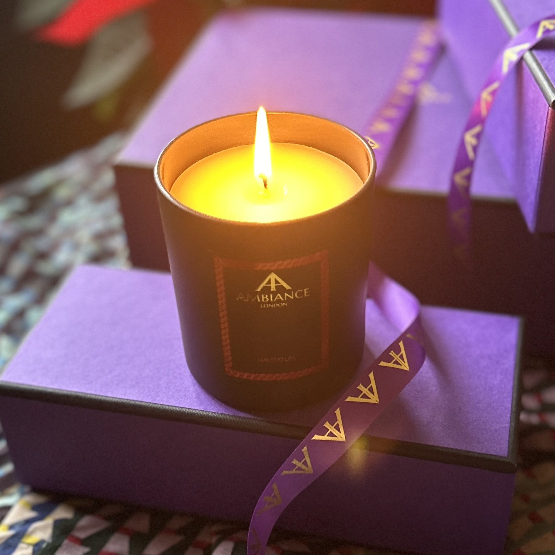 A lit Ancienne Ambiance London Navitatum Candle sits elegantly amongst a selection of purple gift boxes.