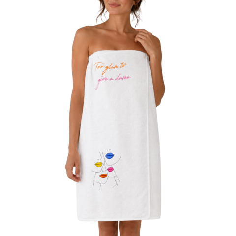 A woman is wrapped in a white towelling wrap dress that says "Too glam to give a damn," featuring colourful abstract faces. The mood is playful and confident.