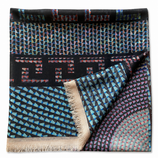 Folded intricate woven pattern wool scarf with concentric circles, geometric borders, and a Greek key design in teal and black. It’s bordered with beige fringe.
