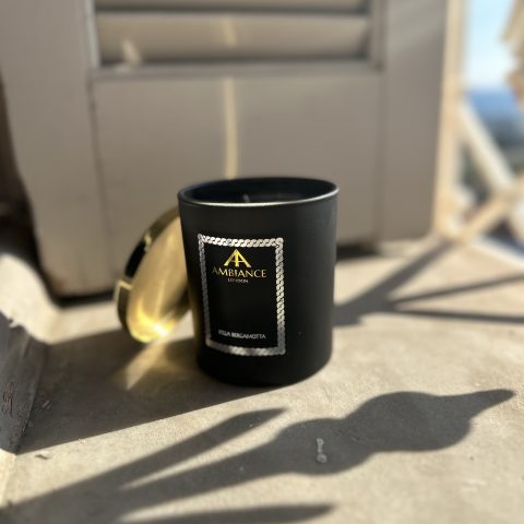 A black glass encased Bella Bergamotta scented candle sits on a window sill. Next to the candle is a golden candle lid and a beige shutter with a blue sky and sea view in the distance.