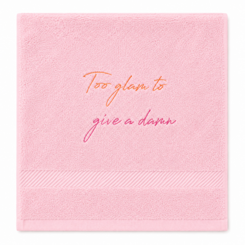 A pink hand towel with the embroidered phrase "Too glam to give a damn" in orange and pink cursive. The design conveys a playful, confident tone.