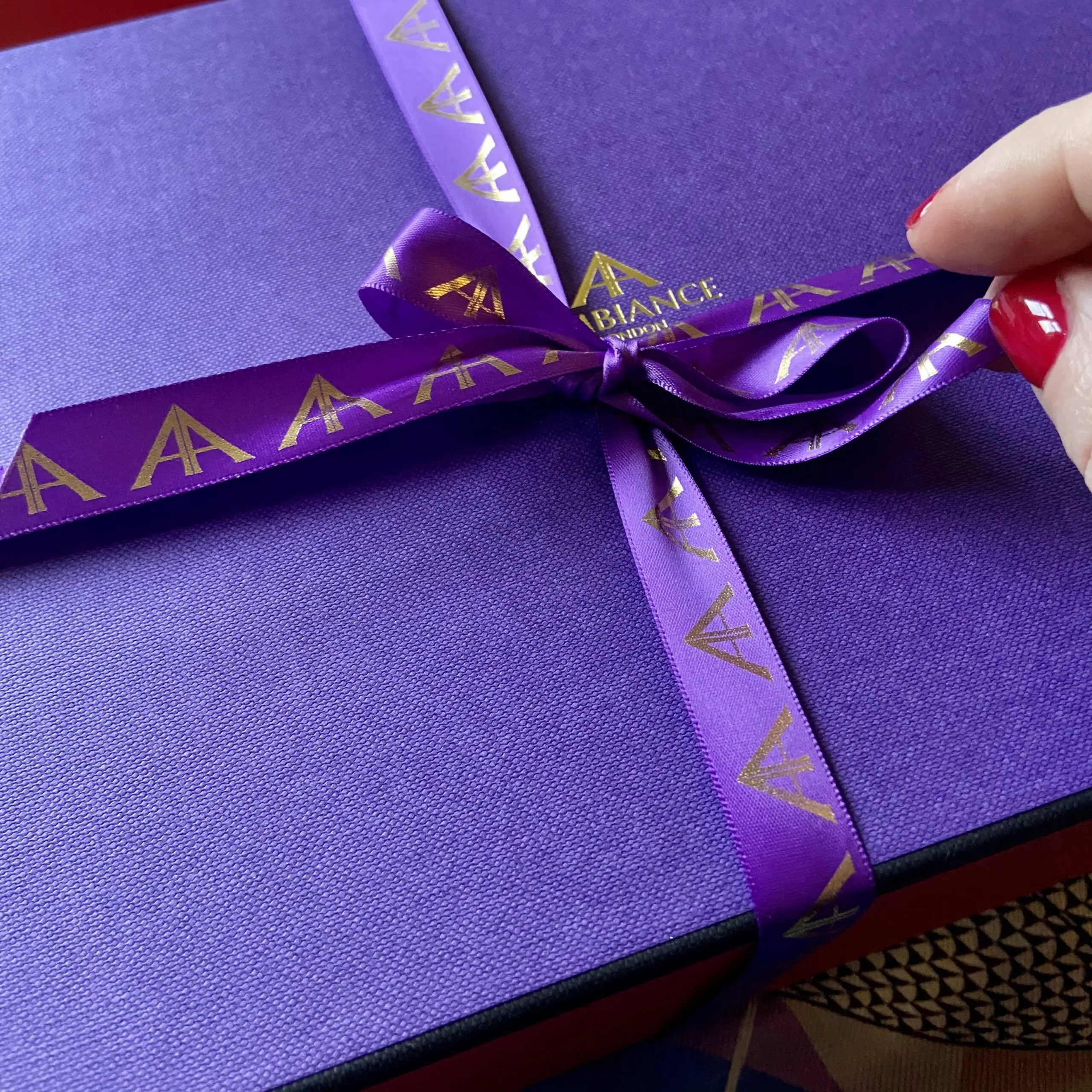 A hand with red nail polish unties a regal purple gift box wrapped with a matching ribbon adorned with gold logos, conveying elegance and anticipation.