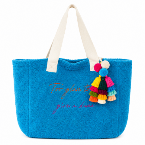 Bright blue towelling tote bag with white cotton handles, featuring the phrase "Too glam to give a damn" embroidered in rainbow colours. Adorned with colourful tassels.