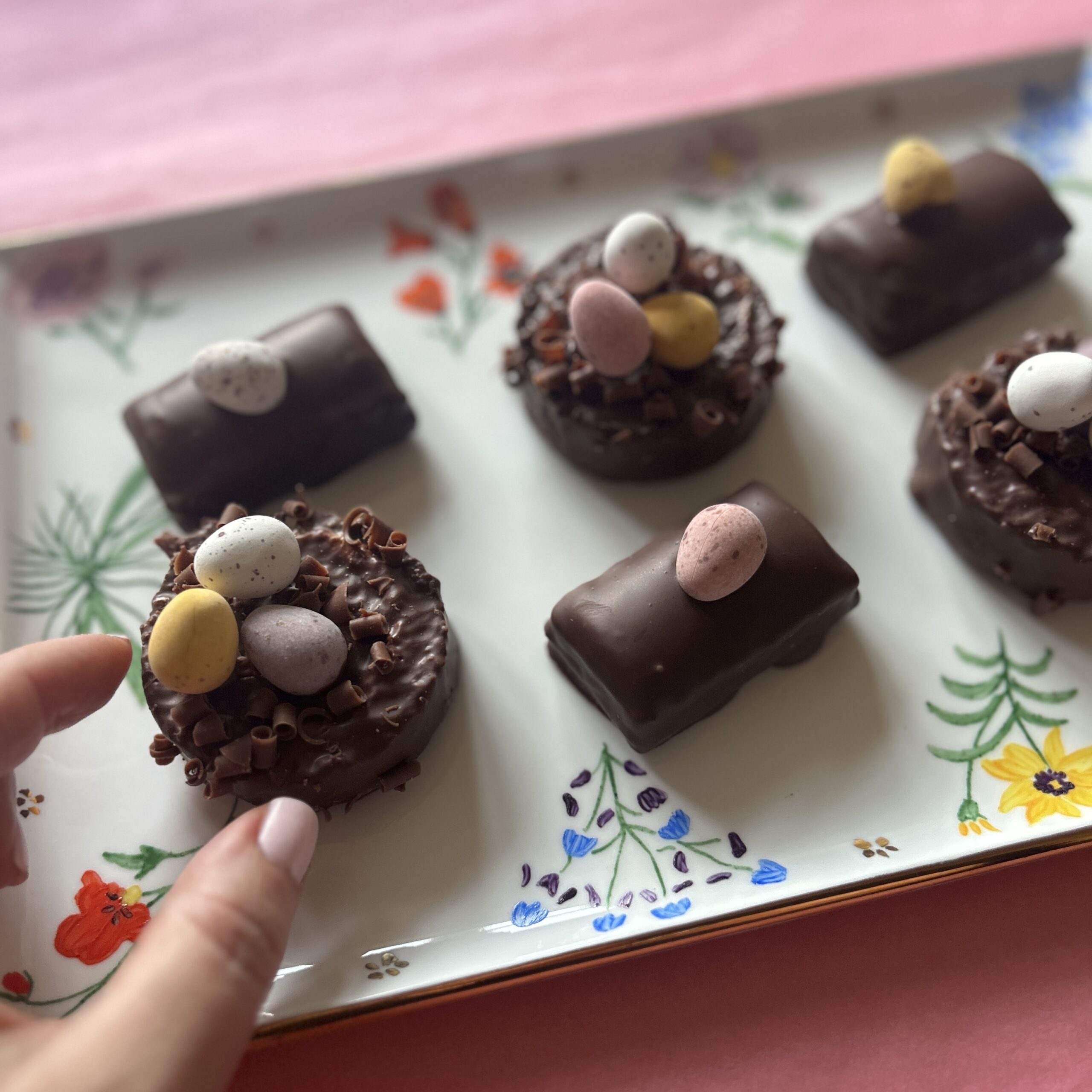 A hand reaches for a chocolate cake decorated with mini eggs on a white floral porcelain platter.