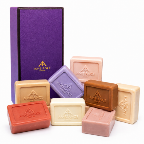 A collection of eight colourful Ancienne Ambiance London soap bars with embossed logos surrounds a tall, elegant purple box. The arrangement conveys luxury and variety.
