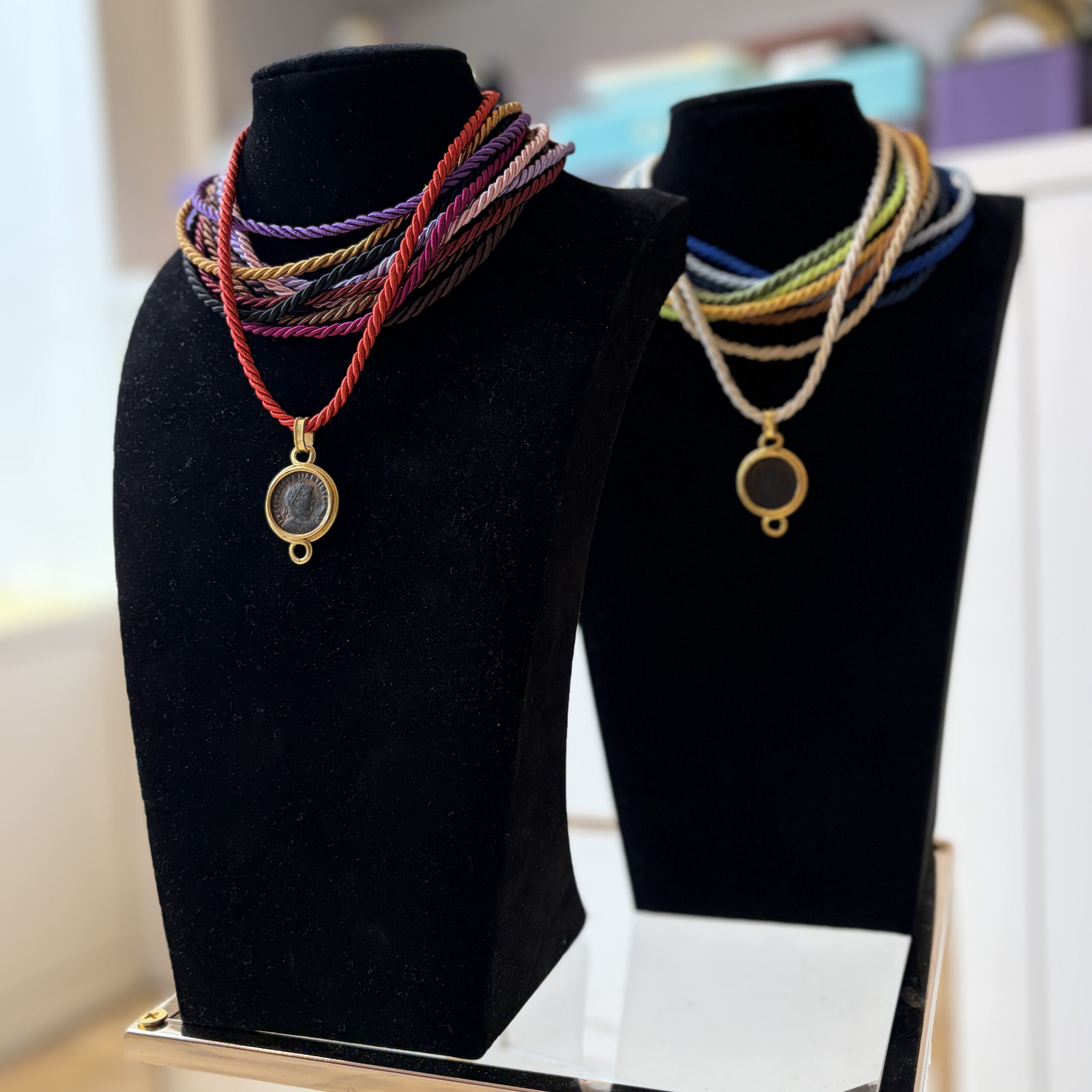 Two black velvet necklace displays hold colourful, twisted cord necklaces with Ancient Roman bronze coin pendants set with 21K gold. The vibrant coluors and gold accents add a stylish touch.