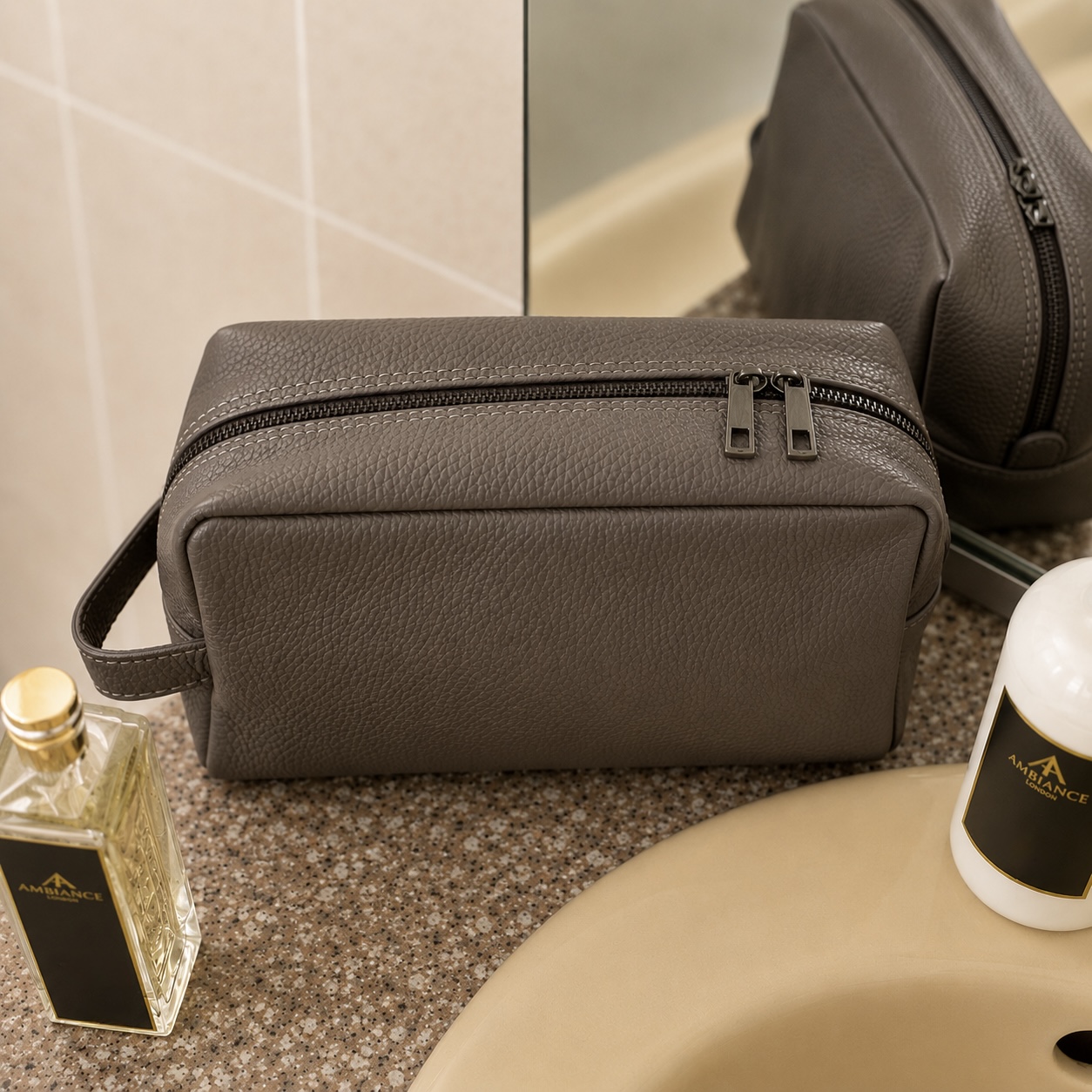 A grey leather toiletry bag with a zipper sits on a speckled countertop next to a gold-capped perfume bottle, creating a neat and sophisticated setting.