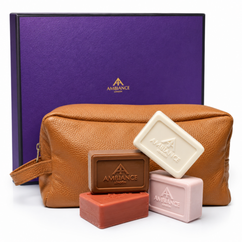 A stylish leather toiletry bag is accompanied by four bars of soap in cream, brown, red and pink, in front of a luxurious purple Ancienne Ambiance London box.