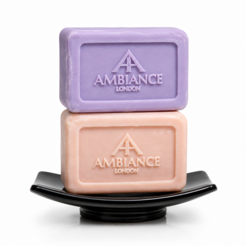 Two bars of soap are stacked on a black dish: a purple bar on top and a peach one beneath, both embossed with "AMBIANCE LONDON," on a white background.