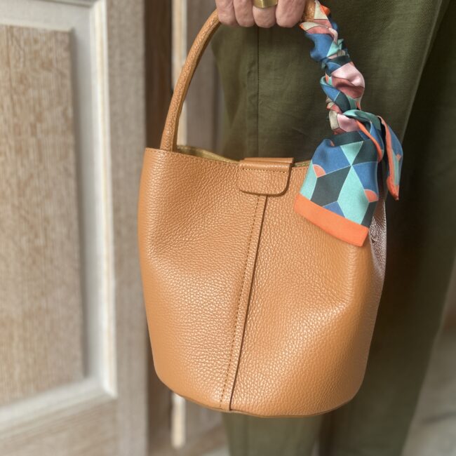 A lady holds a Tan Leather Dalu Bucket Bag by the top handle. The bag is adorned with a twilly scarf.