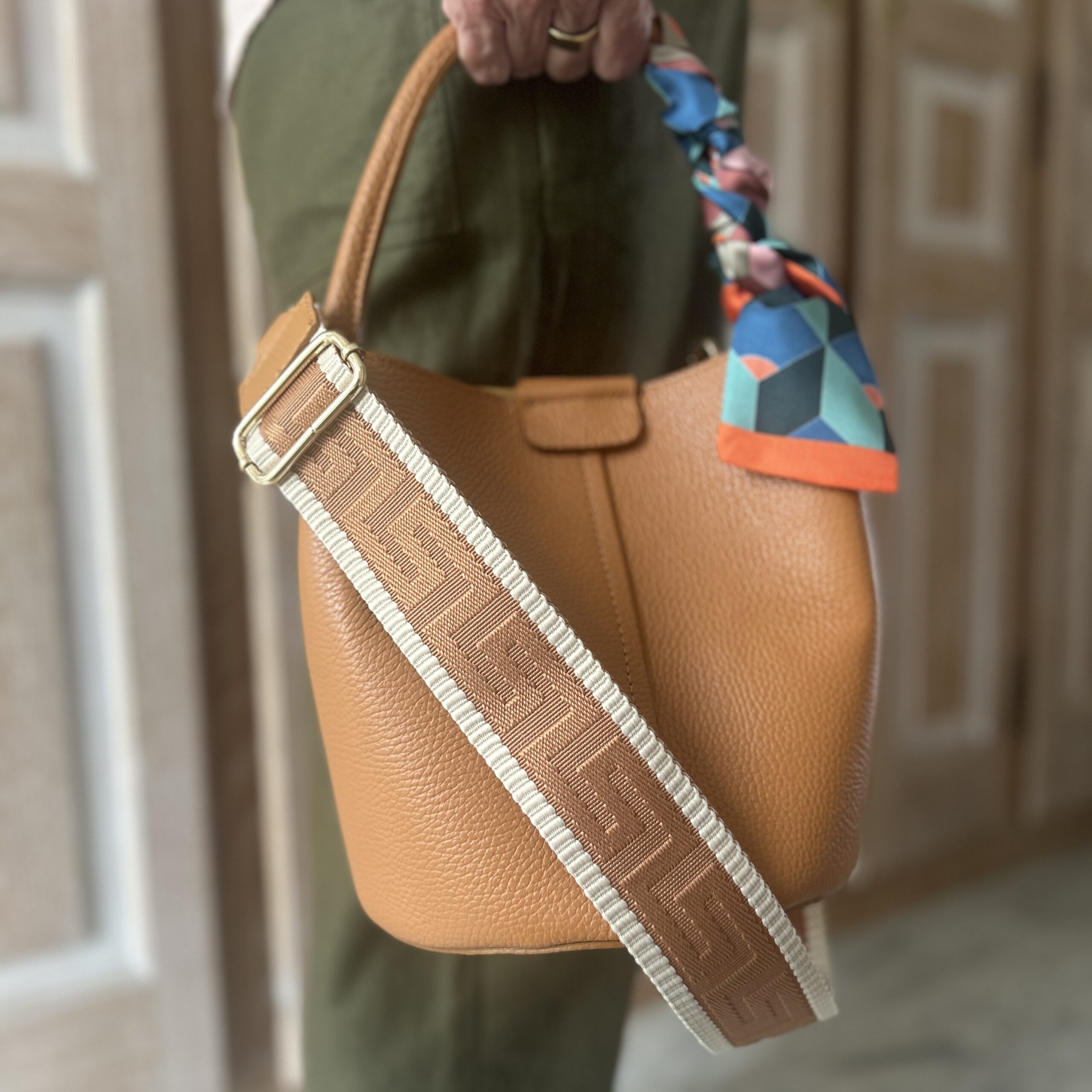 A lady holds a Tan Leather Dalu Bucket Bag by the top handle. The bag is adorned with a twilly scarf and shoulder strap.