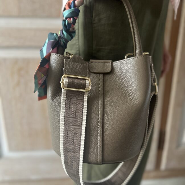 A lady holds a Taupe Leather Dalu Bucket Bag with twilly scarf on the top handle. A shoulder strap drapes over the bag.