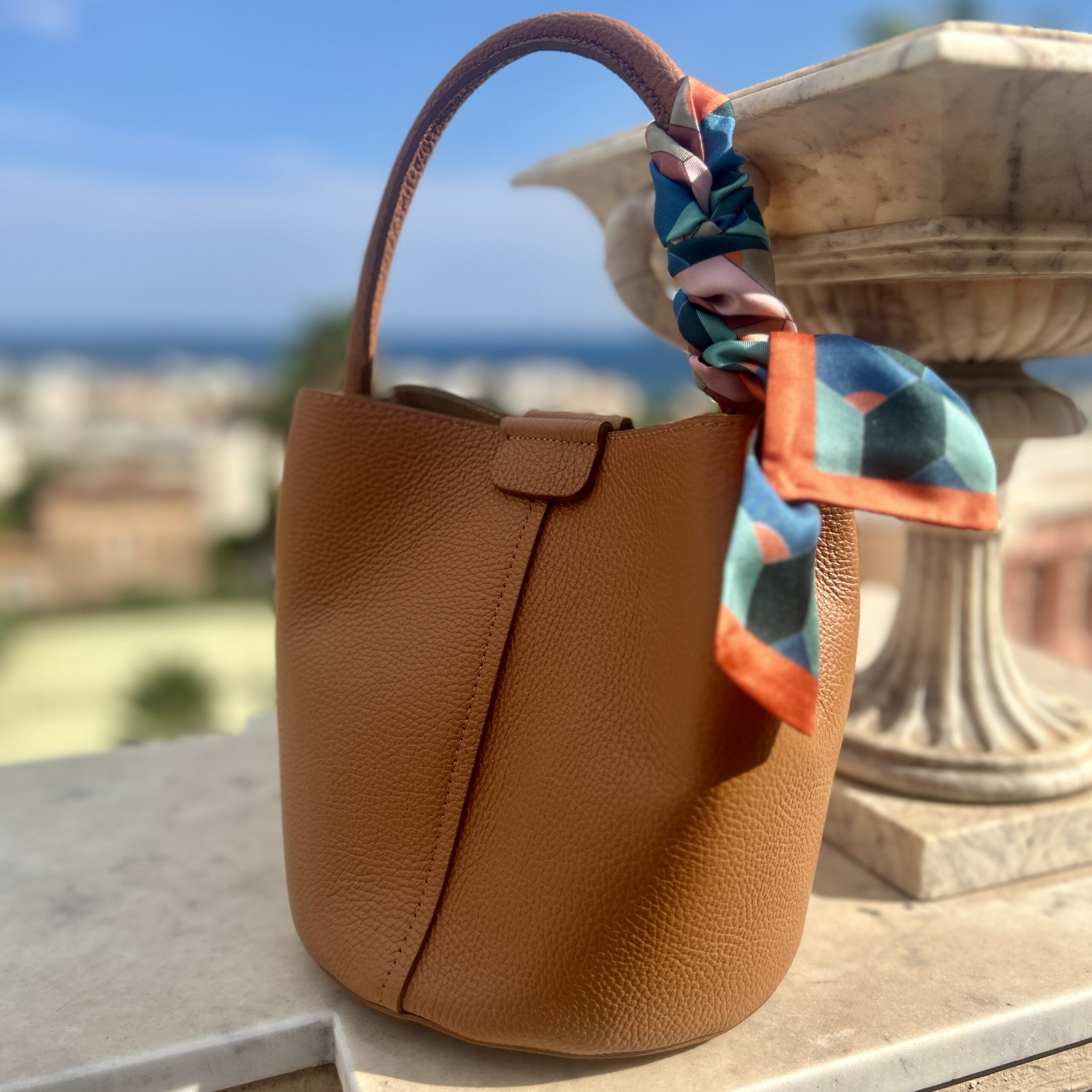 Tan leather handbag with a colourful geometric silk twilly scarf tied to the handle, placed on a stone ledge. Softly blurred sea view background under a clear blue sky.