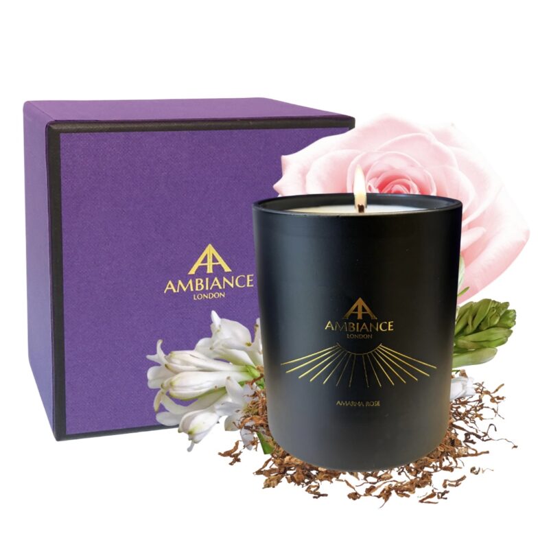 A black candle with a lit wick and gold “Ancienne Ambiance London” logo, surrounded by white tuberose and a pink rose. A purple box stands in the background. Elegant and soothing.