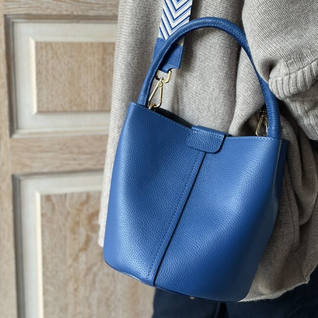 A vibrant blue pebble leather handbag, featuring gold hardware and a patterned strap, hangs against a beige knitted sweater.