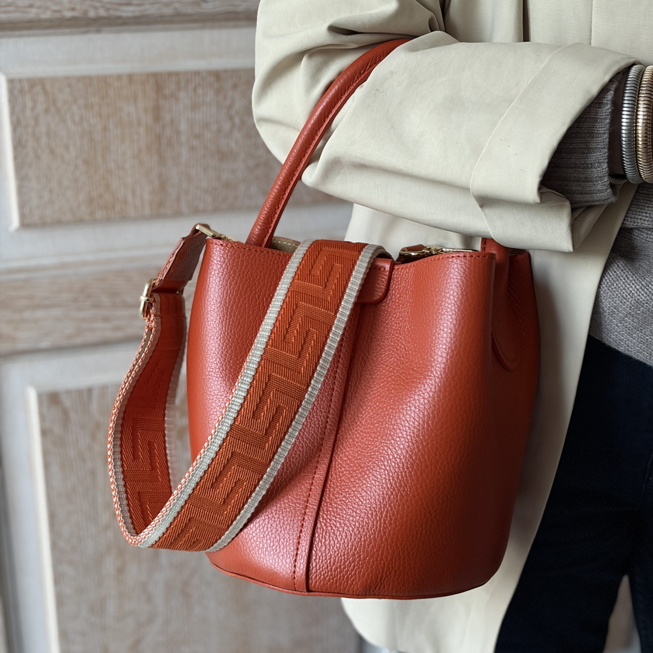 Person holding an orange leather bucket bag with a patterned strap. The outfit includes a beige coat and a grey sweater, conveying a stylish tone.