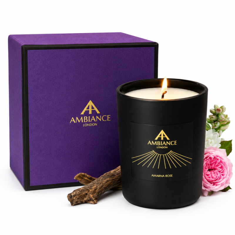 A lit black candle labeled "Ancienne Ambiance London Amarna Rose" is beside a purple box with gold lettering. Pink roses, tuberose and brown wood add elegance and warmth.