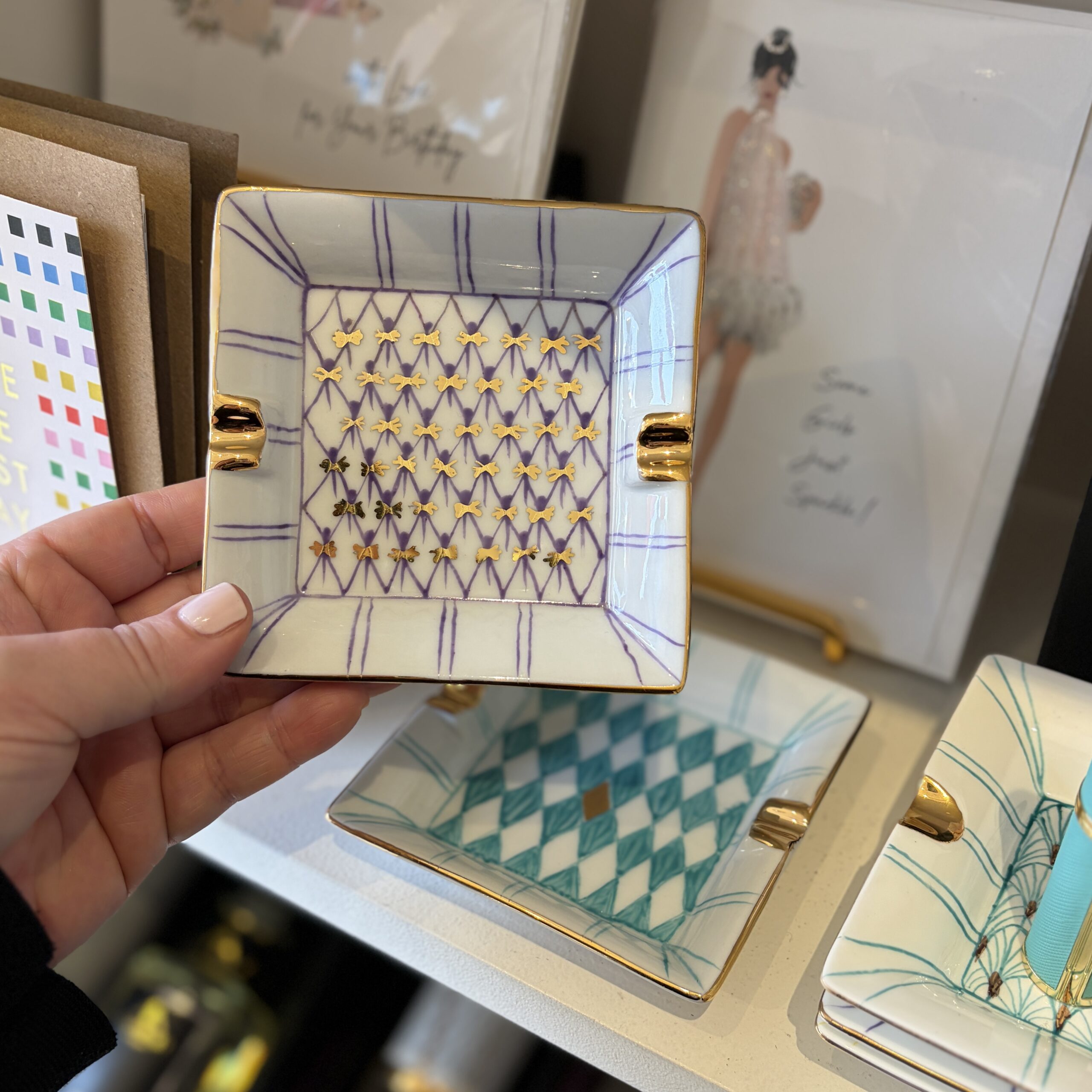 A hand holds a purple square Porcelain Tray with gold plating above a turquoise square porcelain tray on a white shelf.