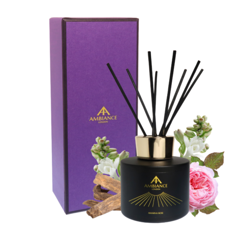 Black scented reed diffuser with gold "Ancienne Ambiance London" logo, labeled "Amarna Rose." Behind it is a purple gift box. Surrounding flowers and wooden accents add elegance and freshness.