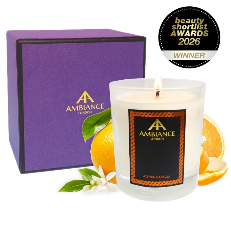 A scented candle labeled "Ancienne Ambiance London Assyria Blossom" sits lit beside a purple box. Fresh oranges and blossoms accent the scene. The candle is an award winner, noted by a "Beauty Shortlist Awards 2026 Winner" badge.