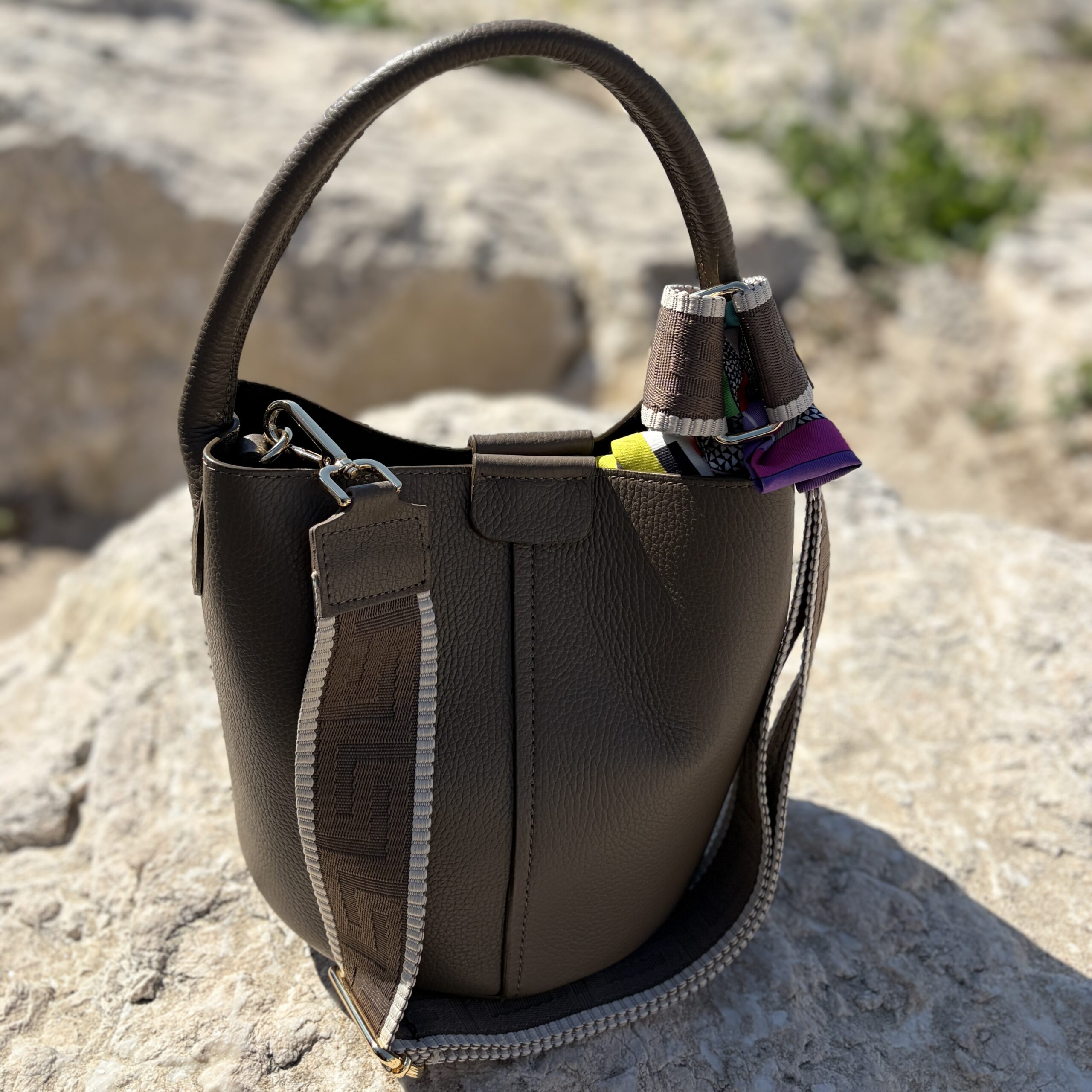 A taupe leather bucket bag with a handle and colourful twilly detail sits on sunlit rocks by the beach, conveying a casual elegance.