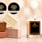 Ancienne Ambiance London products displayed elegantly on a marble surface, featuring soap bars, a lit assyria blossom candle and a bottle of goddess beauty oil. Three beauty awards badges are shown above.