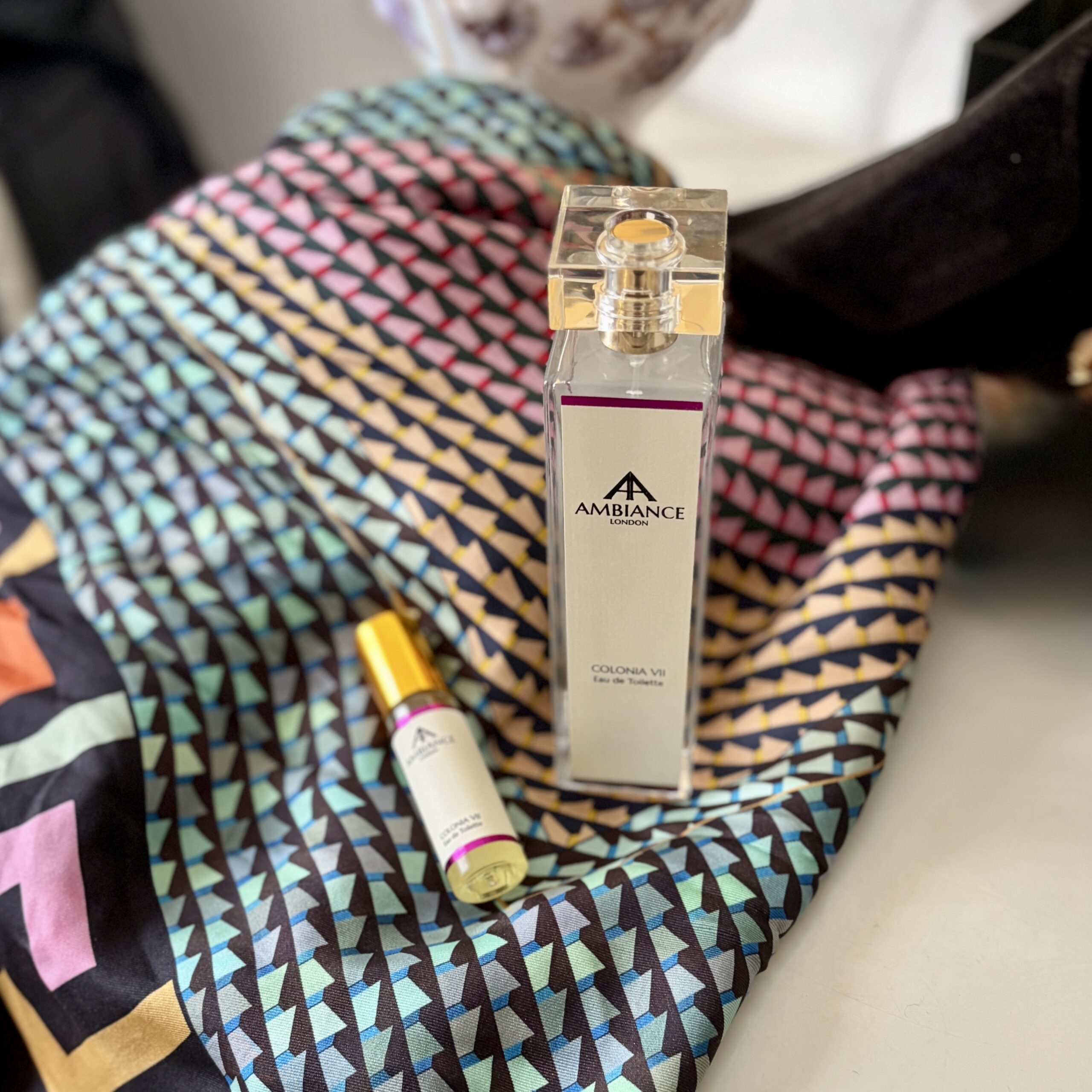 Perfume bottles labeled "Ancienne Ambiance London eau de toilette" on a colourful, geometric-patterned silk scarf. The scene conveys elegance and sophistication.