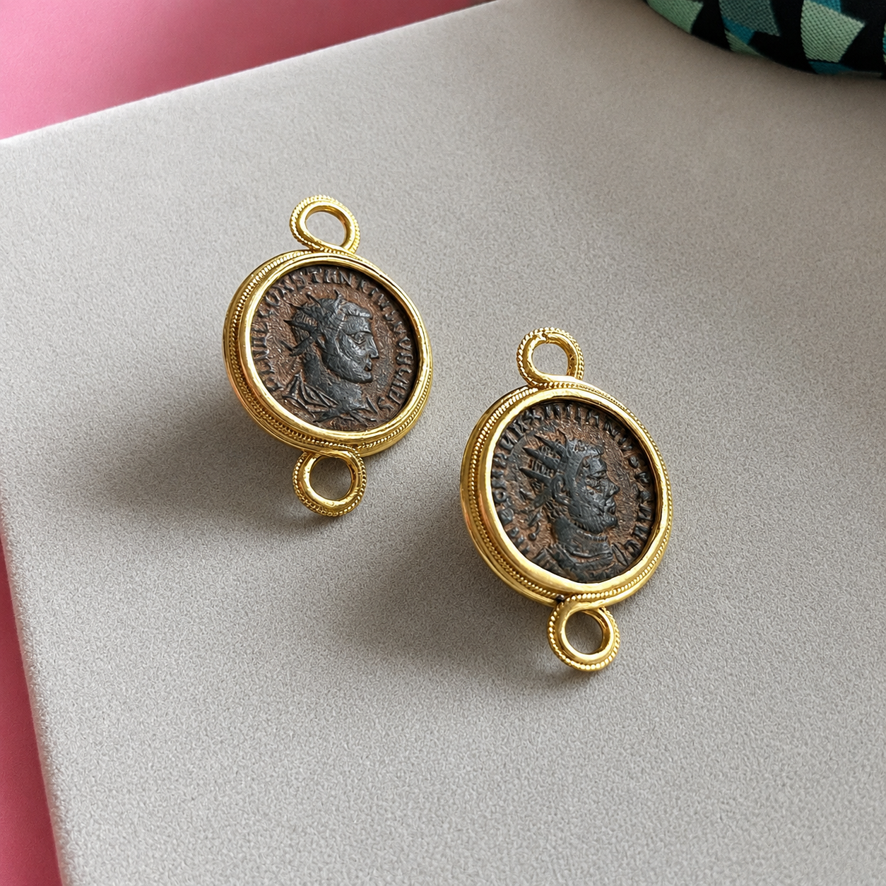 Two rustic coins set in ornate gold frames, featuring a profile of a figure, placed on a soft gray surface with a pink background corner.