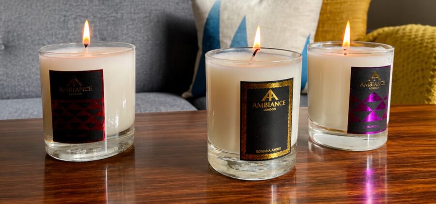 Three lit candles in glass holders sit on a wooden table in a cosy living room. The background features a grey couch with colourful pillows and framed art.