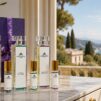Elegant perfume bottles with golden caps and a purple gift box are displayed on a marble table. The backdrop features a serene coastal view with trees and blue sky.