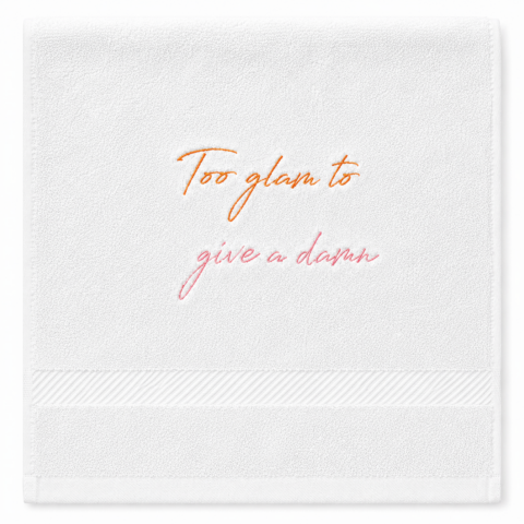 White hand towel with embroidered text, "Too glam to give a damn" in orange and pink cursive. The design conveys confidence and elegance.