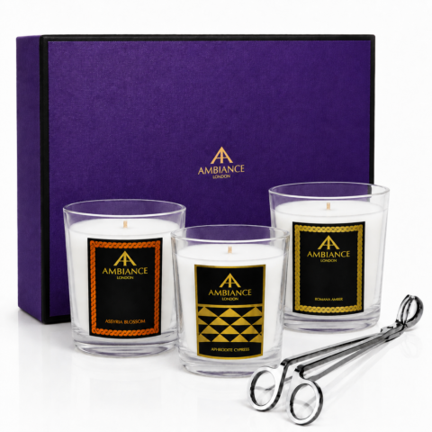 Three candles with decorative labels are placed before a purple gift box with "Ancienne Ambiance London" branding. Silver wick trimmer lays nearby, evoking elegance.