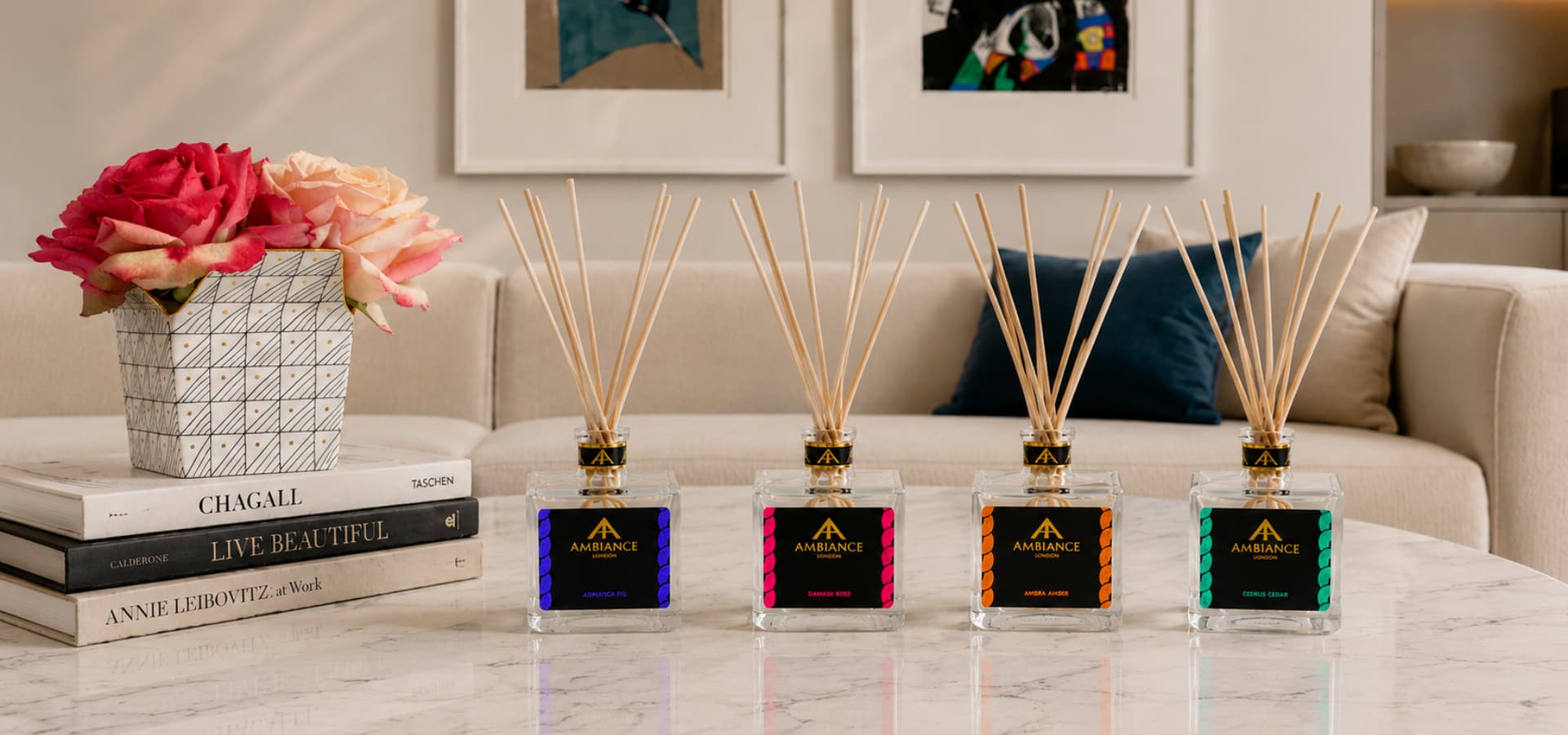 A row of four elegant diffuser bottles with coloured labels and reed sticks sits on a marble table beside a vase of pink roses and stacked books in a chic living room.