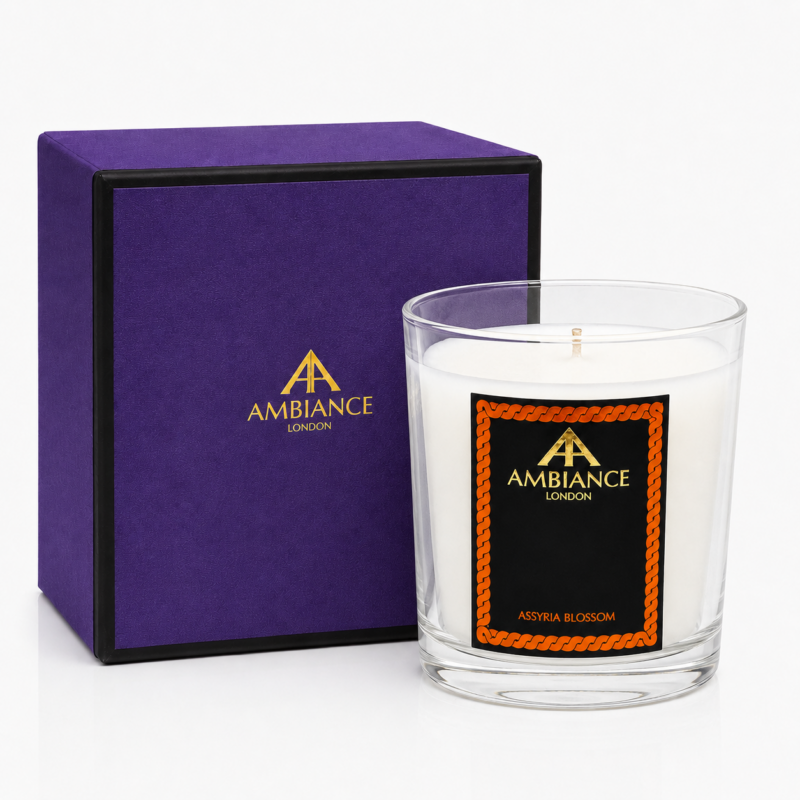 Elegant Ancienne Ambiance London candle in a glass holder with a black and orange label, labeled "Assyria Blossom." Purple box beside it, creating a luxurious feel.