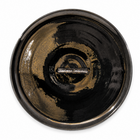 A round ceramic plate with glossy black and bronze swirls, featuring a metallic rectangular "Ancienne Ambiance" logo at the centre. The design is modern and artistic.