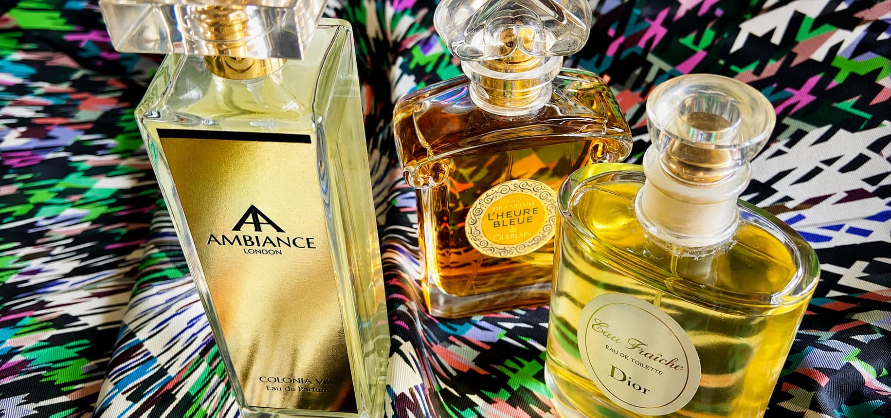 Three elegant perfume bottles—Ancienne Ambiance Colonia, L'Heure Bleue, and Dior—are displayed on a vibrant, multicoloured fabric, conveying luxury and sophistication.