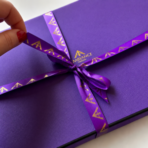 A hand with red nail polish pulls a purple satin ribbon from a purple gift box. The ribbon features gold "A" logos and "Ancienne Ambiance London" branding.