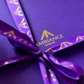 A vibrant purple gift box tied with a matching purple ribbon featuring golden "Ancienne Ambiance London" logo. A hand with red nails gently holds the ribbon. The tone is elegant.