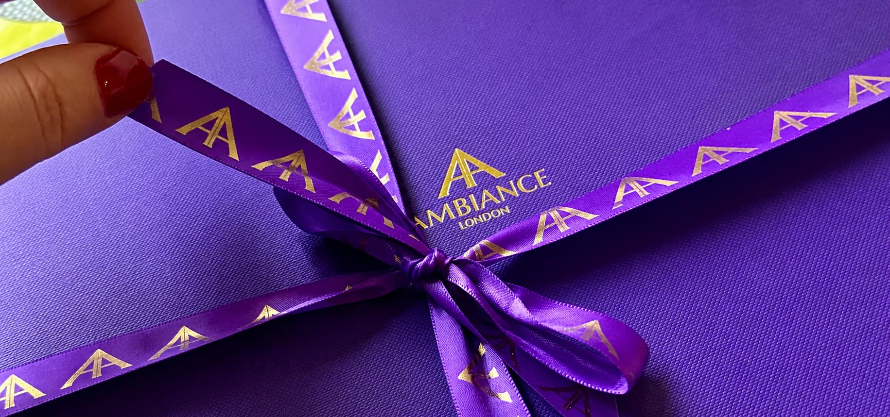 A vibrant purple gift box tied with a matching purple ribbon featuring golden "Ancienne Ambiance London" logo. A hand with red nails gently holds the ribbon. The tone is elegant.
