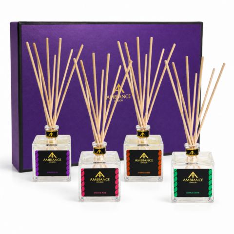 Four elegantly Ancienne Ambiance London reed diffusers are placed in front of a purple gift box. The diffusers feature square glass bottles with long natural reeds.