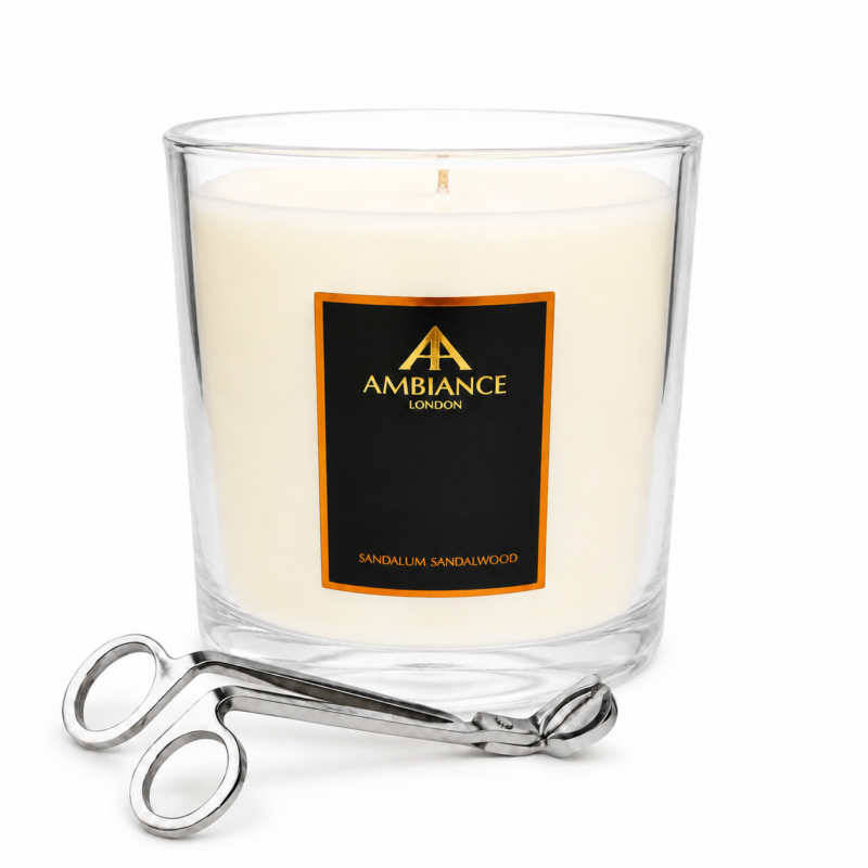 A sandalwood-scented candle in a large clear glass jar with a black and gold "Ancienne Ambiance London" label. Beside it are silver wick trimmers, creating an elegant setting.