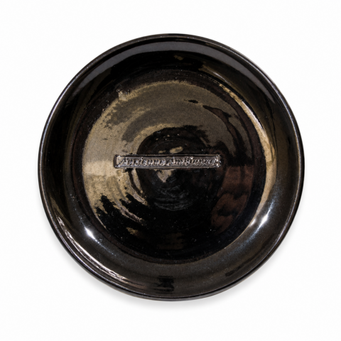 A black ceramic plate with a glossy finish, featuring the engraving "Ancienne Ambiance" at the centre. The plate has a reflective, elegant appearance.