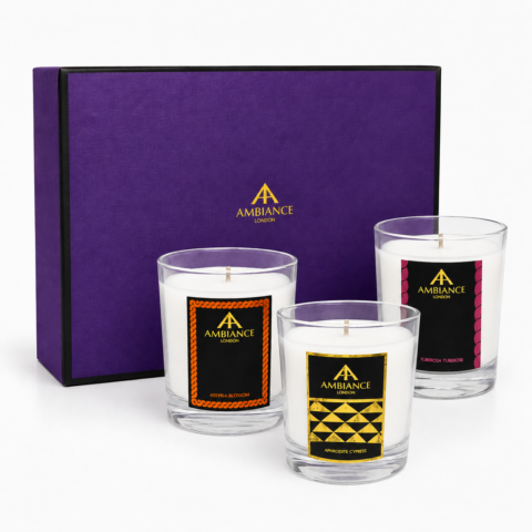 Three Ancienne Ambiance London candles in glass holders with colourful labels are in front of a purple gift box. The labels feature geometric patterns.