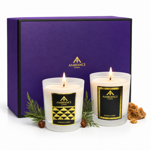 Two lit candles in glass holders labeled "Ambiance London" with elegant black and gold designs. They sit in front of a purple gift box, exuding a luxurious and serene atmosphere.