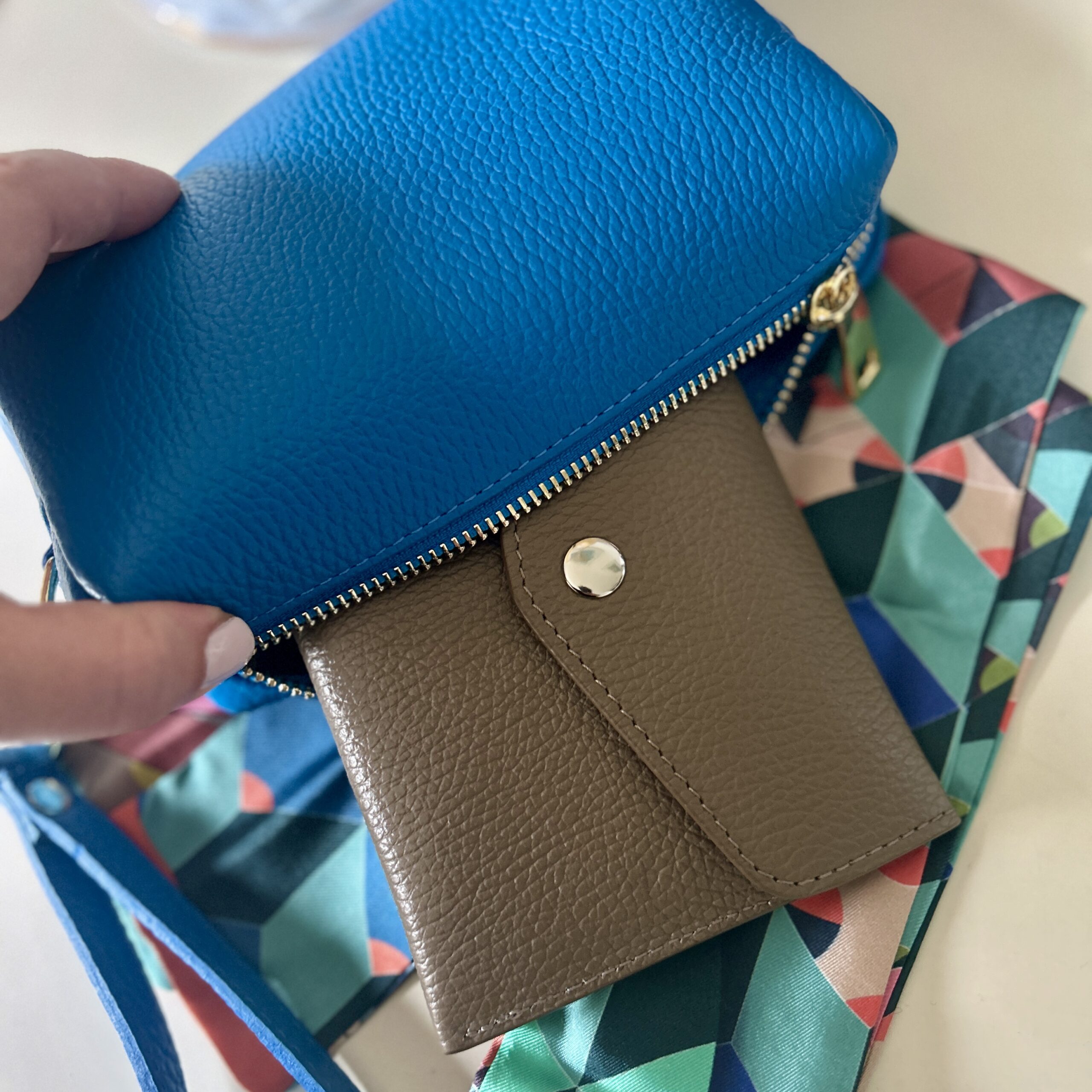A hand holds a bright blue leather pouch with a gold zipper, revealing a taupe textured wallet. Below is a colourful geometric-patterned silk scarf.