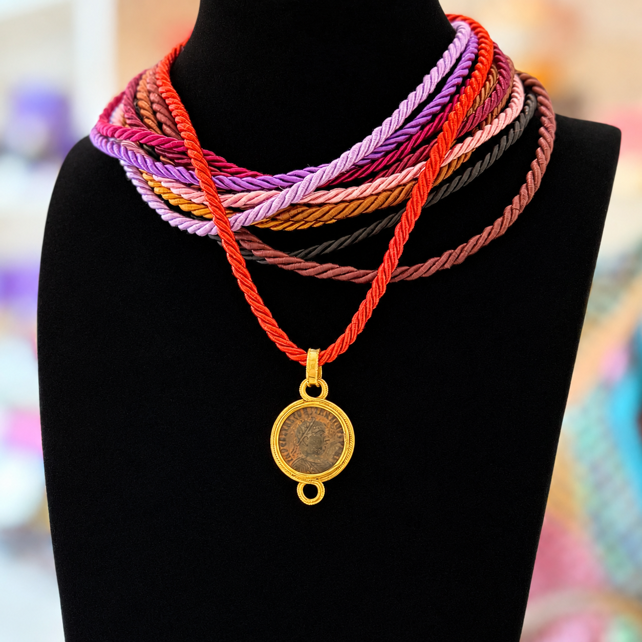 A black mannequin displays colourful, intertwined cord necklaces in red, purple, and pink hues, adorned with a gold pendant featuring an ancient Roman coin.
